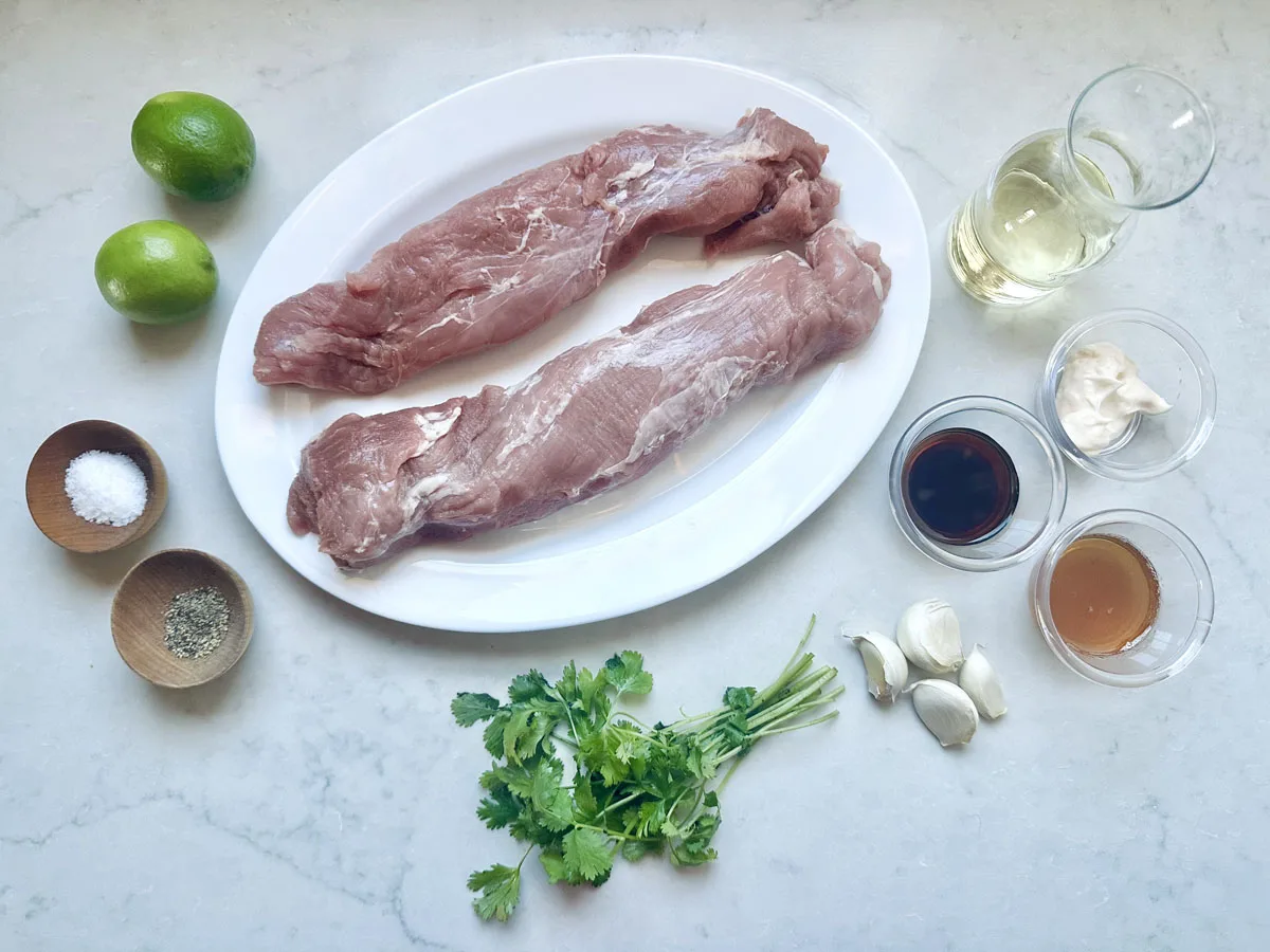 Two raw pork tenderloins on a white oval plate, surrounded by limes, a glass of white wine, salt, pepper, cilantro, garlic cloves, soy sauce, mayonnaise, and a brown liquid in small bowls on a light countertop.