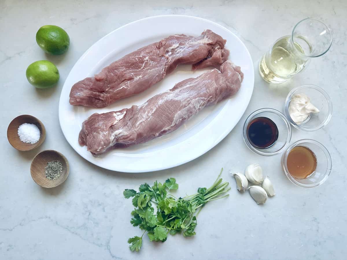 Two raw pork tenderloins on a white oval plate, surrounded by limes, a glass of white wine, salt, pepper, cilantro, garlic cloves, soy sauce, mayonnaise, and a brown liquid in small bowls on a light countertop.