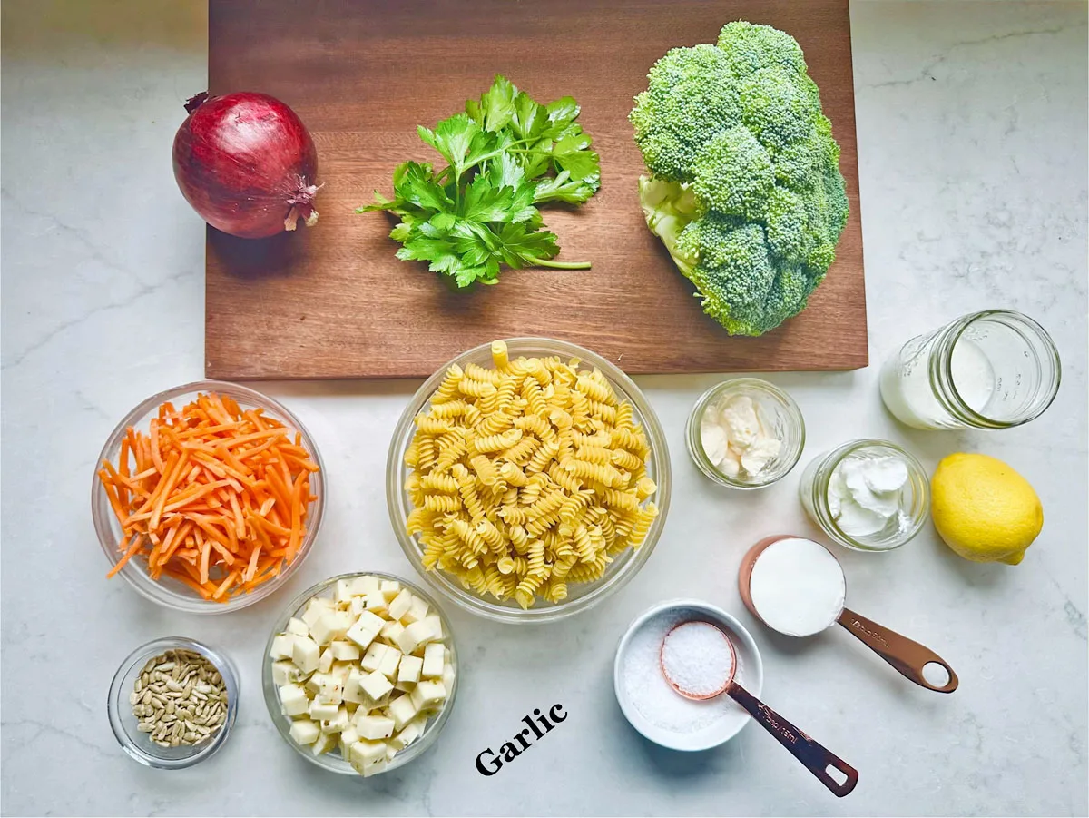 A variety of ingredients are arranged on a counter for Pasta Salad with Broccoli and Carrots: a red onion, parsley, broccoli, shredded carrots, cubed PepperJack Cheese, rotini pasta, garlic, sunflower seeds, lemon, salt, sour cream, oil, and a wooden cutting board.
