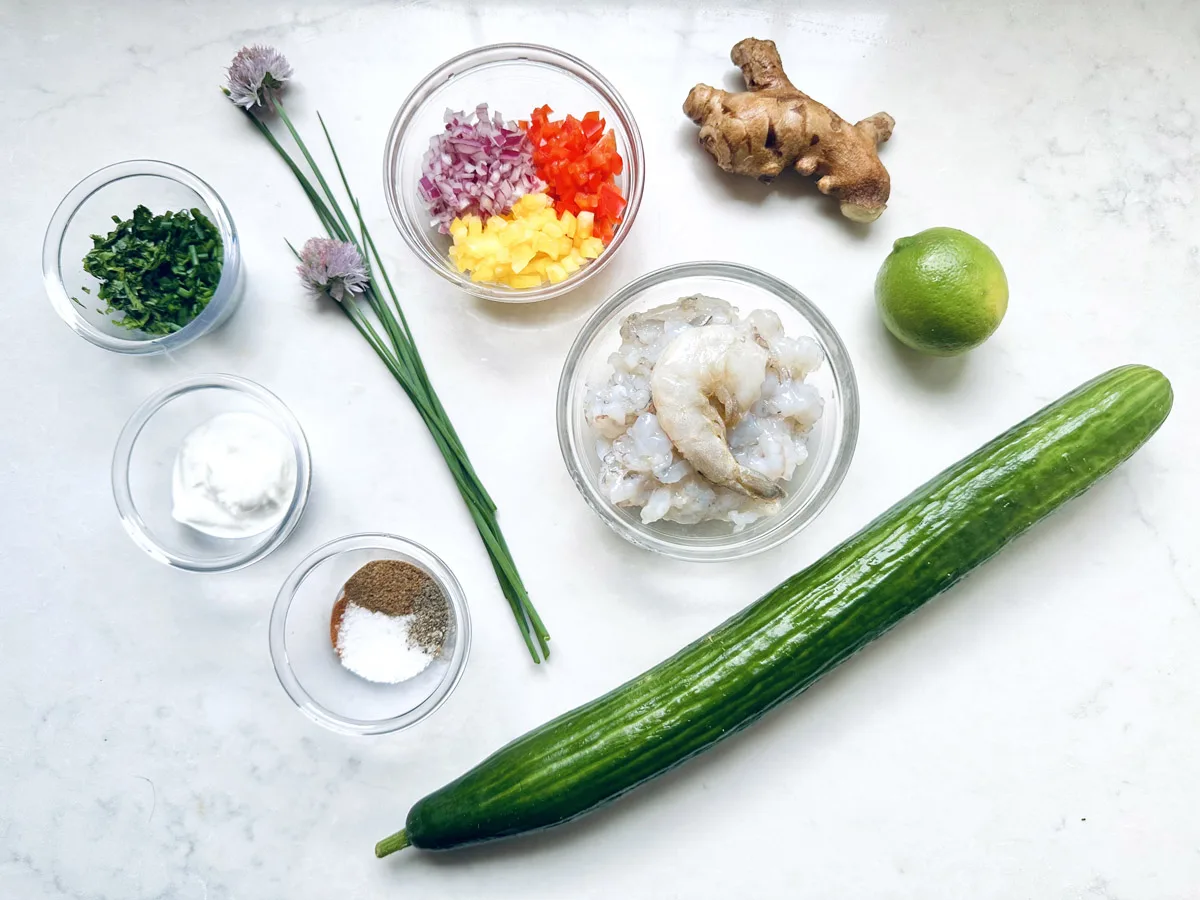 Top-down view of cooking ingredients on a white surface, including cucumber, lime, ginger, shrimp, chopped herbs, chopped red onion and bell pepper, yogurt, and small bowls of salt and pepper.