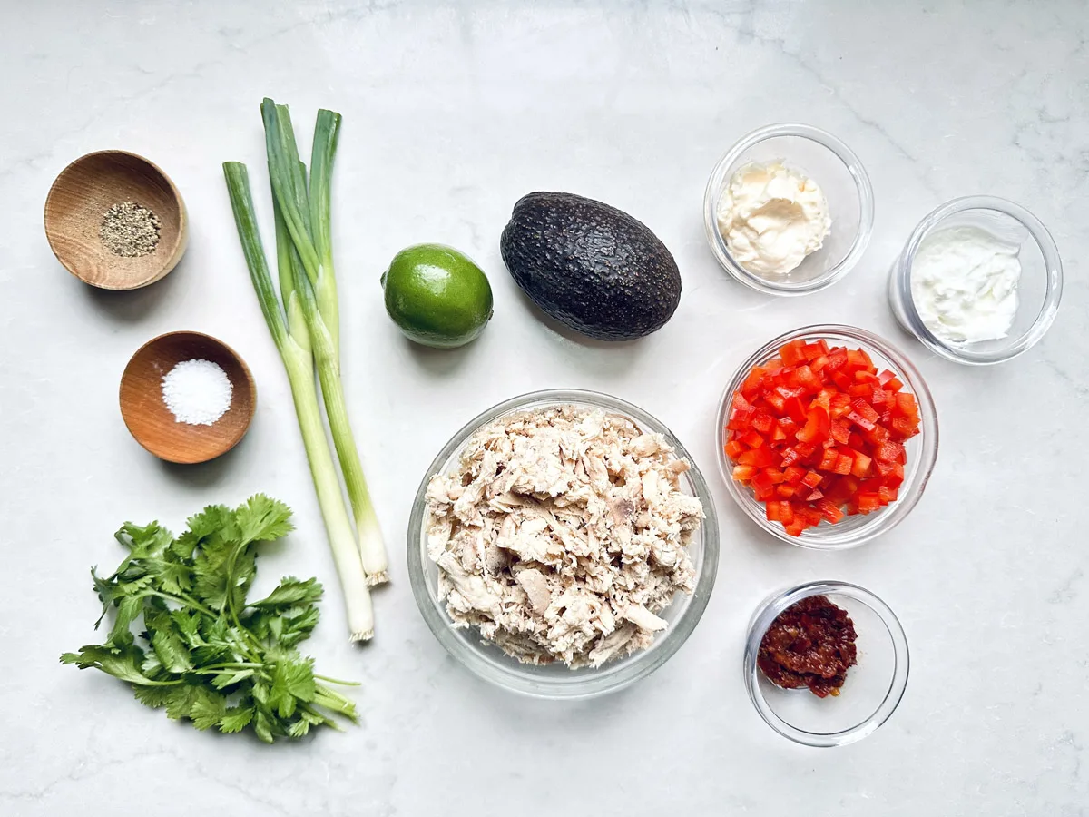Ingredients laid out on a white surface for Spicy Chicken Salad with Chipotle and Avocado: chopped cooked chicken, diced red bell pepper, avocado, green onions, cilantro, lime, seasonings, mayonnaise, yogurt, and chili paste in small bowls.