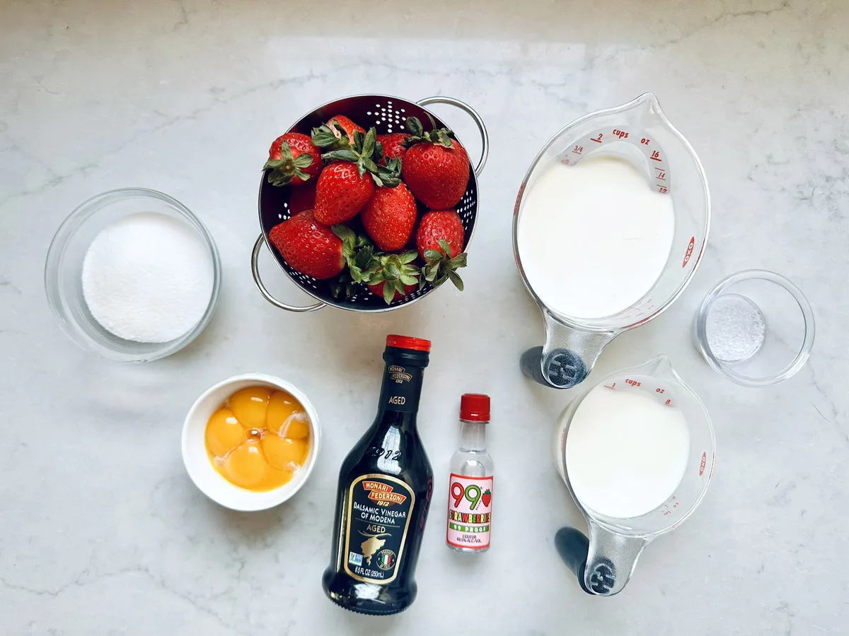 A top-down view of ingredients for Strawberry-Balsamic Ice Cream on a light surface, including strawberries, granulated sugar, egg yolks, balsamic vinegar, vanilla extract, milk, heavy cream, and a pinch of salt in measuring cups and bowls.