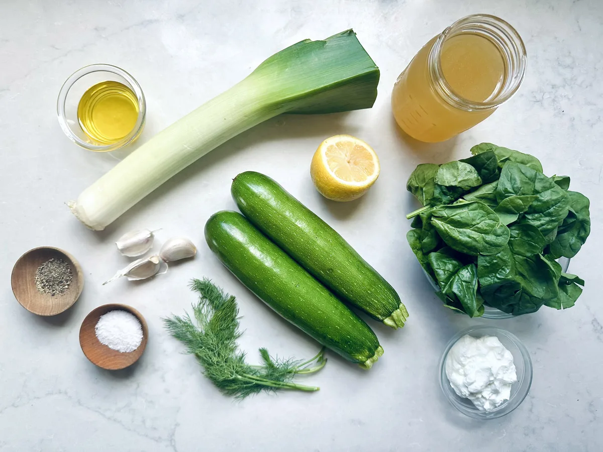A flat lay of ingredients for Zucchini Soup with Dill and Greek Yogurt on a white surface, featuring a leek, two zucchinis, fresh spinach, lemon half, dill, garlic cloves, olive oil, yogurt, vegetable broth, salt, and pepper in small bowls.