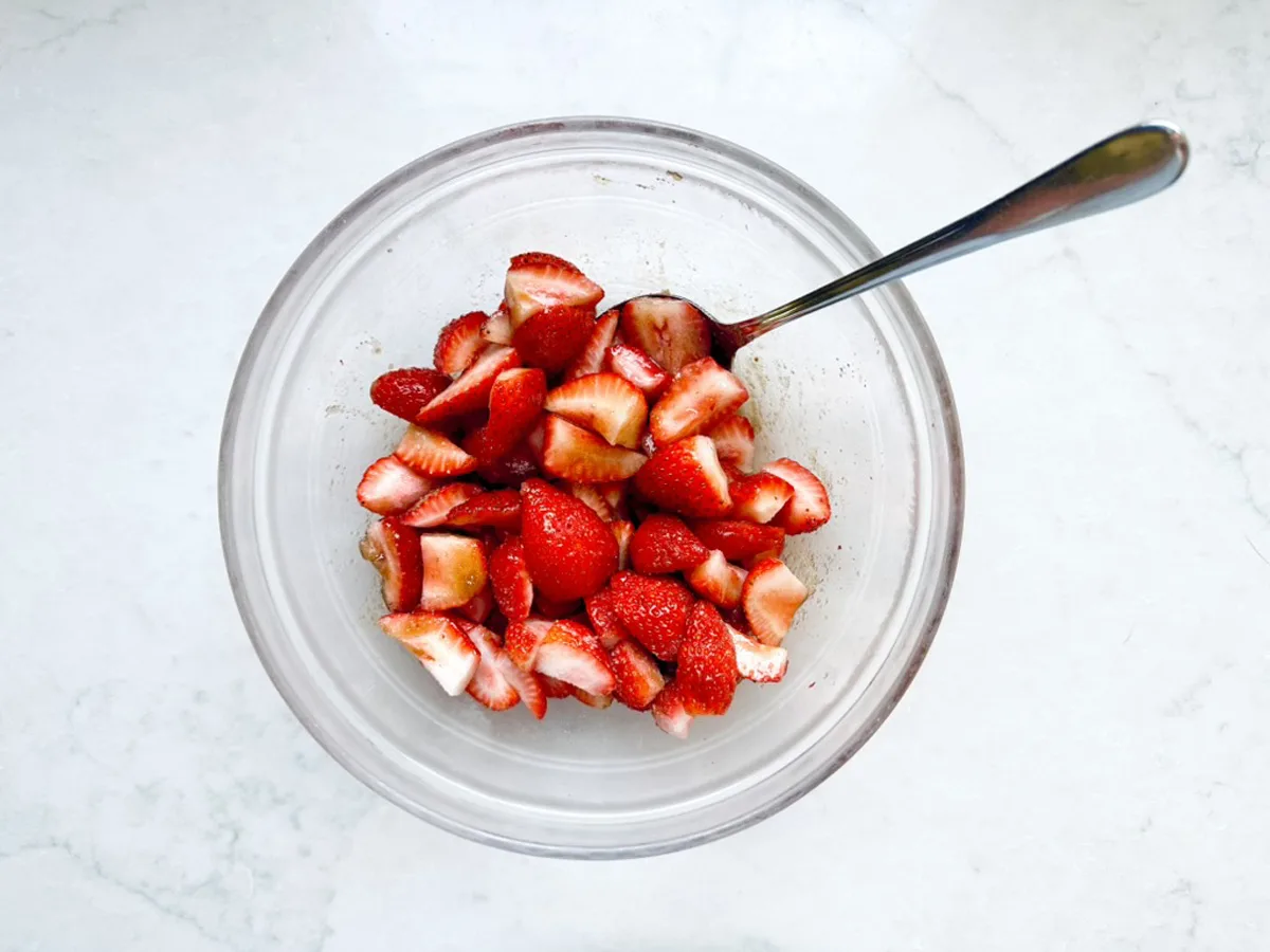 A clear glass bowl filled with sliced strawberries and a metal spoon resting inside, placed on a white marble surface&mdash;perfect for pairing with homemade Strawberry-Balsamic Ice Cream.
