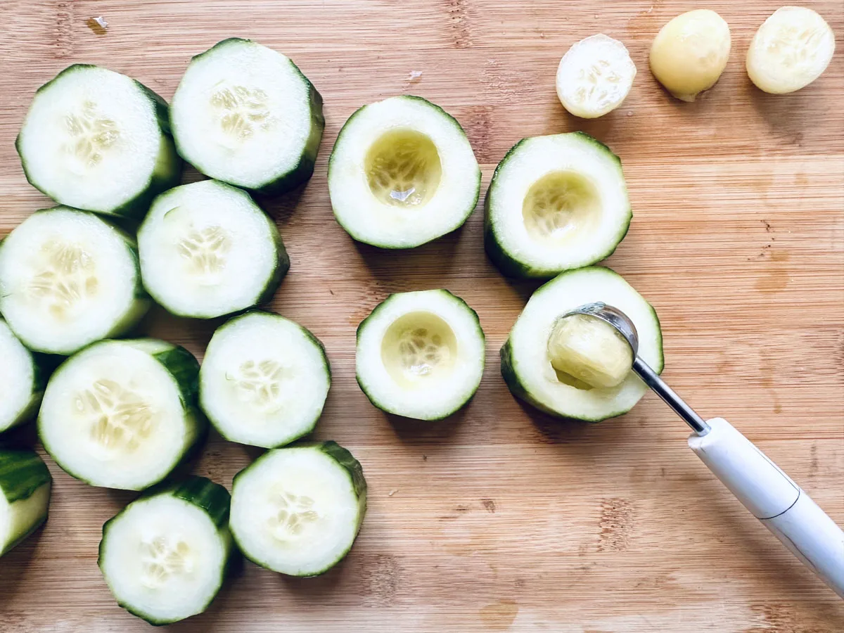 Sliced cucumbers on a wooden cutting board, with some pieces hollowed out using a melon baller tool; scooped cucumber centers are placed to the side.