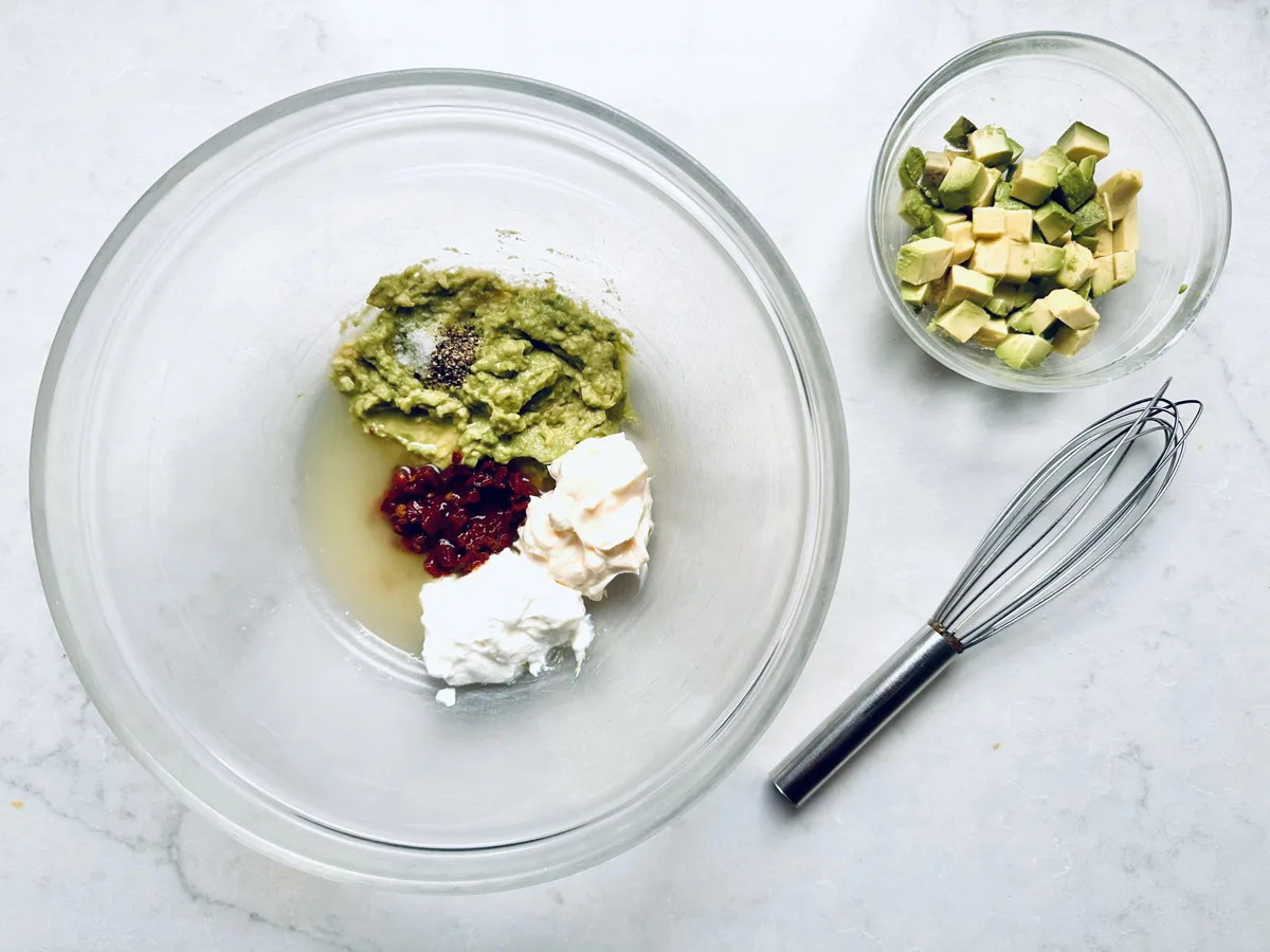 A glass mixing bowl with mashed avocado, yogurt, mayonnaise, chopped sun-dried tomatoes, lime juice, salt, and pepper—perfect for making a Spicy Chicken Salad with Chipotle and Avocado—sits next to a whisk and diced avocado on a white surface.
