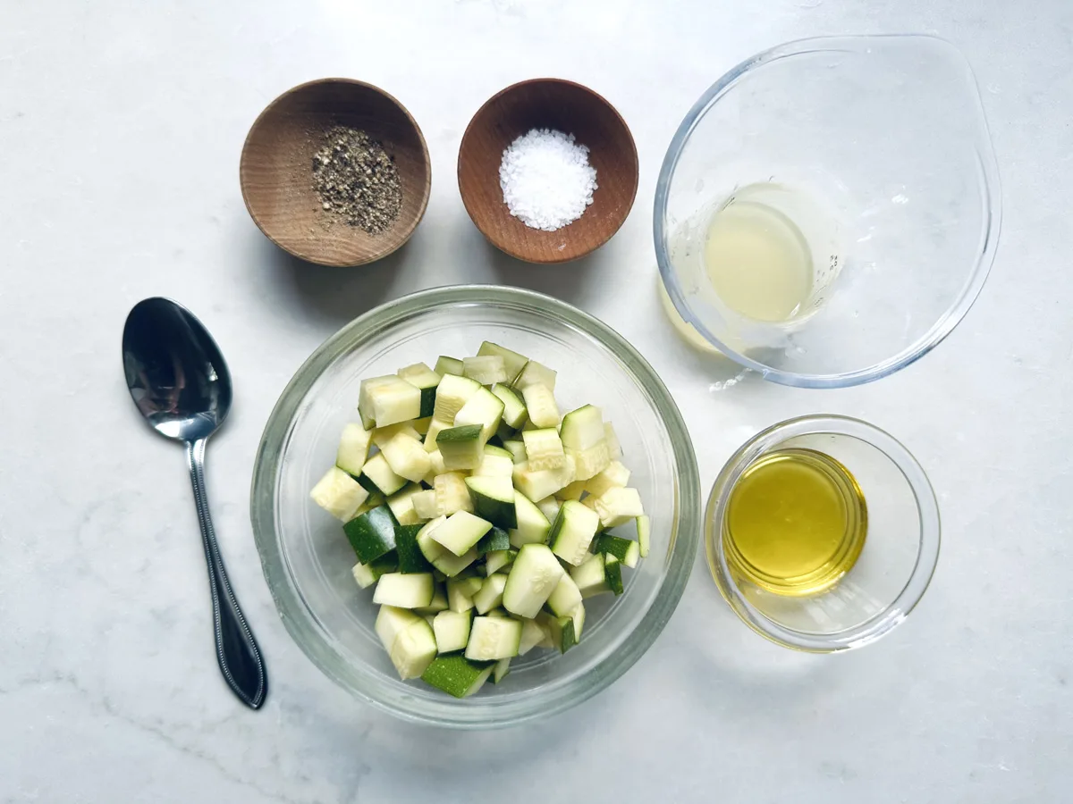 A bowl of chopped zucchini for Zucchini Soup with Dill and Greek Yogurt is surrounded by a spoon, bowls of black pepper and salt, a glass container of lemon juice, and a small bowl of olive oil on a white surface.