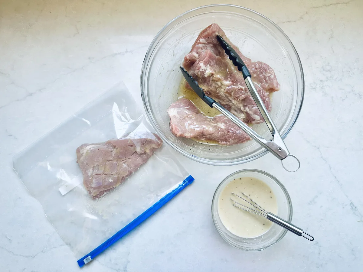 Raw pork chops being marinated in a glass bowl with tongs, one chop in a resealable plastic bag, and a small bowl of marinade with a whisk on a white countertop.
