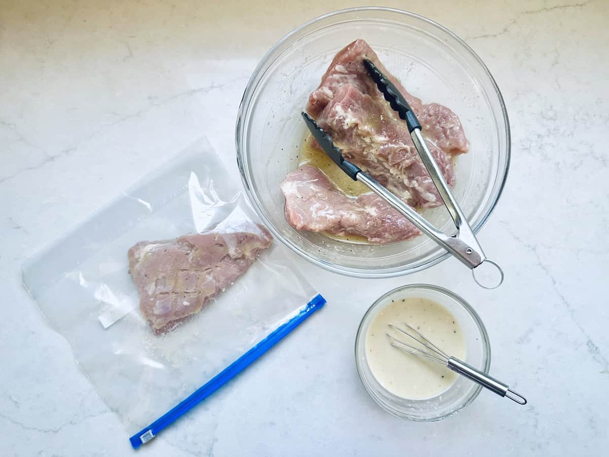 Raw pork chops being marinated in a glass bowl with tongs, one chop in a resealable plastic bag, and a small bowl of marinade with a whisk on a white countertop.