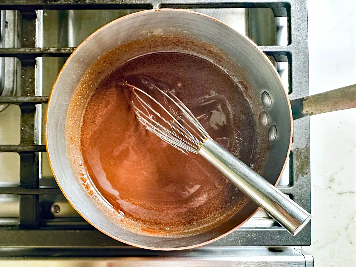 A metal whisk rests in a saucepan filled with a reddish-brown sauce, sitting on a stovetop with visible burner grates—perhaps the beginning of a rich glaze for Chocolate Chipotle Brownies. The scene evokes the art of crafting bold, flavorful desserts.