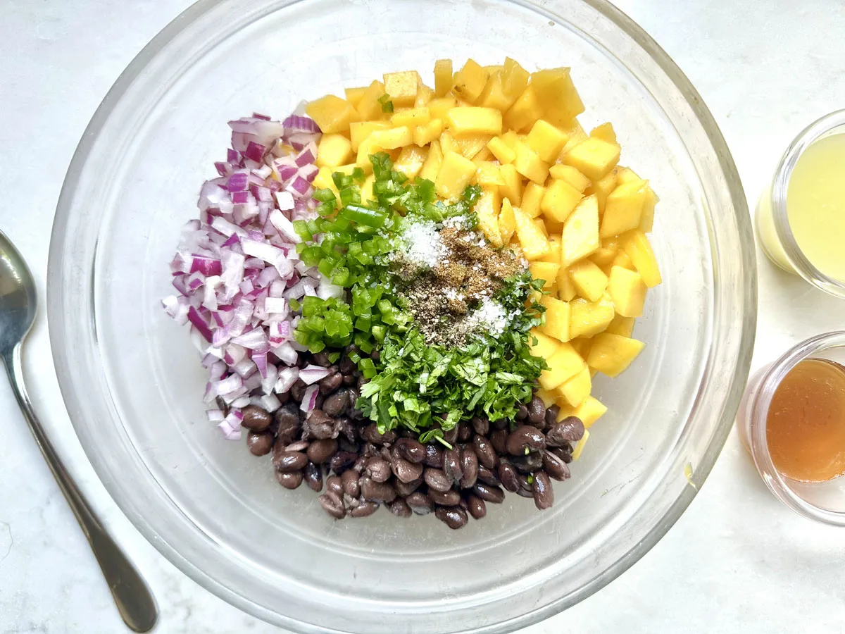 A glass bowl contains diced red onion, mango, jalapeño, black beans, chopped cilantro, and spices. Small bowls of dressing ingredients and a spoon are nearby on a white surface.