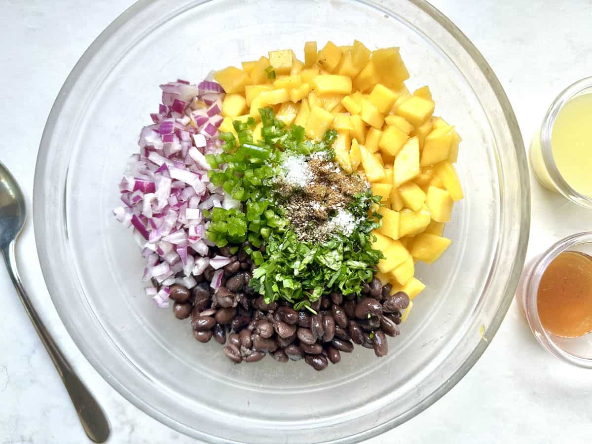 A glass bowl contains diced red onion, mango, jalapeño, black beans, chopped cilantro, and spices. Small bowls of dressing ingredients and a spoon are nearby on a white surface.