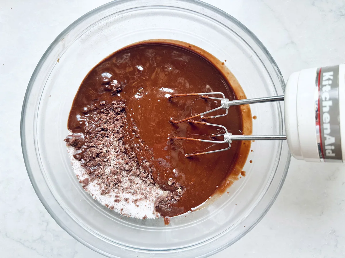 A glass bowl with Chocolate Chipotle Brownies batter being mixed by a white KitchenAid hand mixer on a light countertop. Some dry mix is still visible on one side of the bowl.