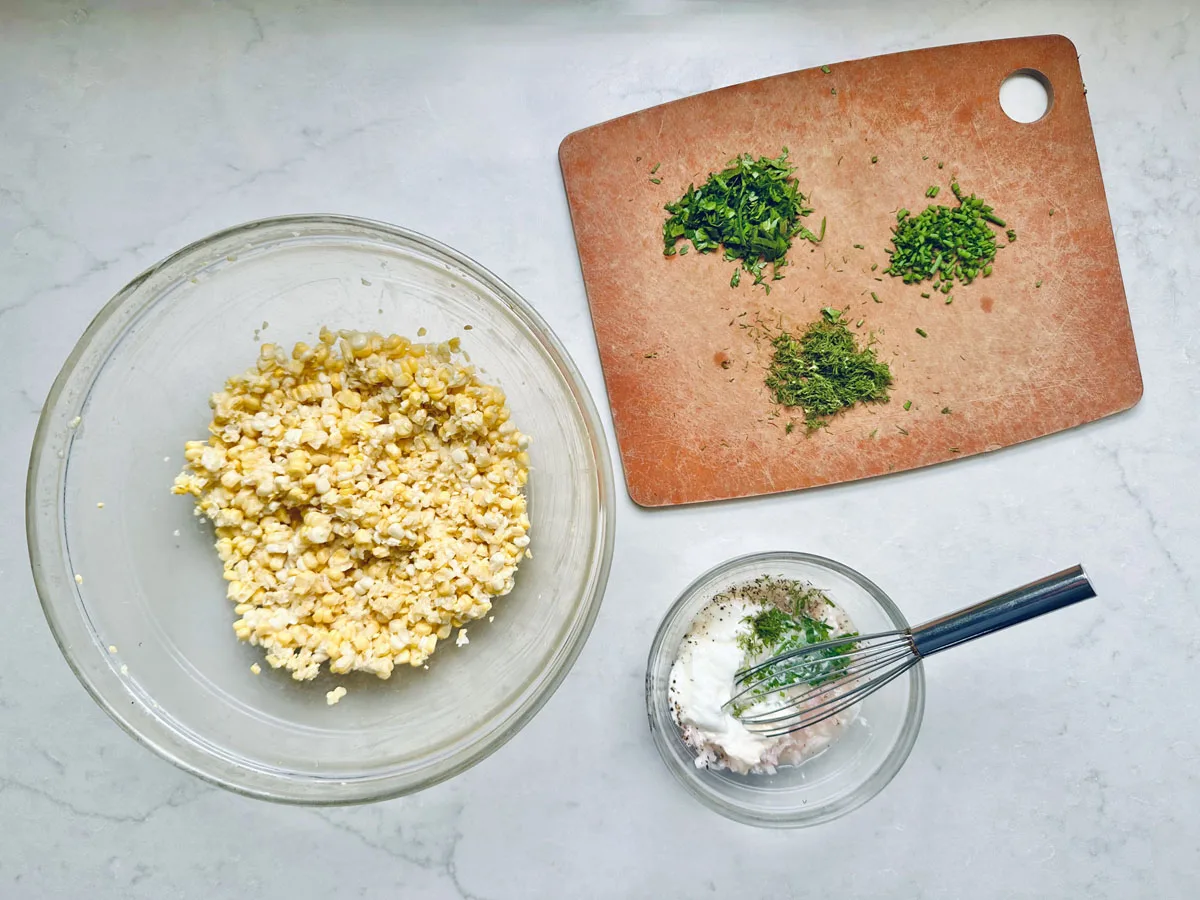 Overhead view of a bowl with chopped hard-boiled eggs, a cutting board with three piles of chopped herbs, and a small bowl with mayonnaise and seasonings being whisked for a Corn Salad with Buttermilk and Herbs on a white countertop.
