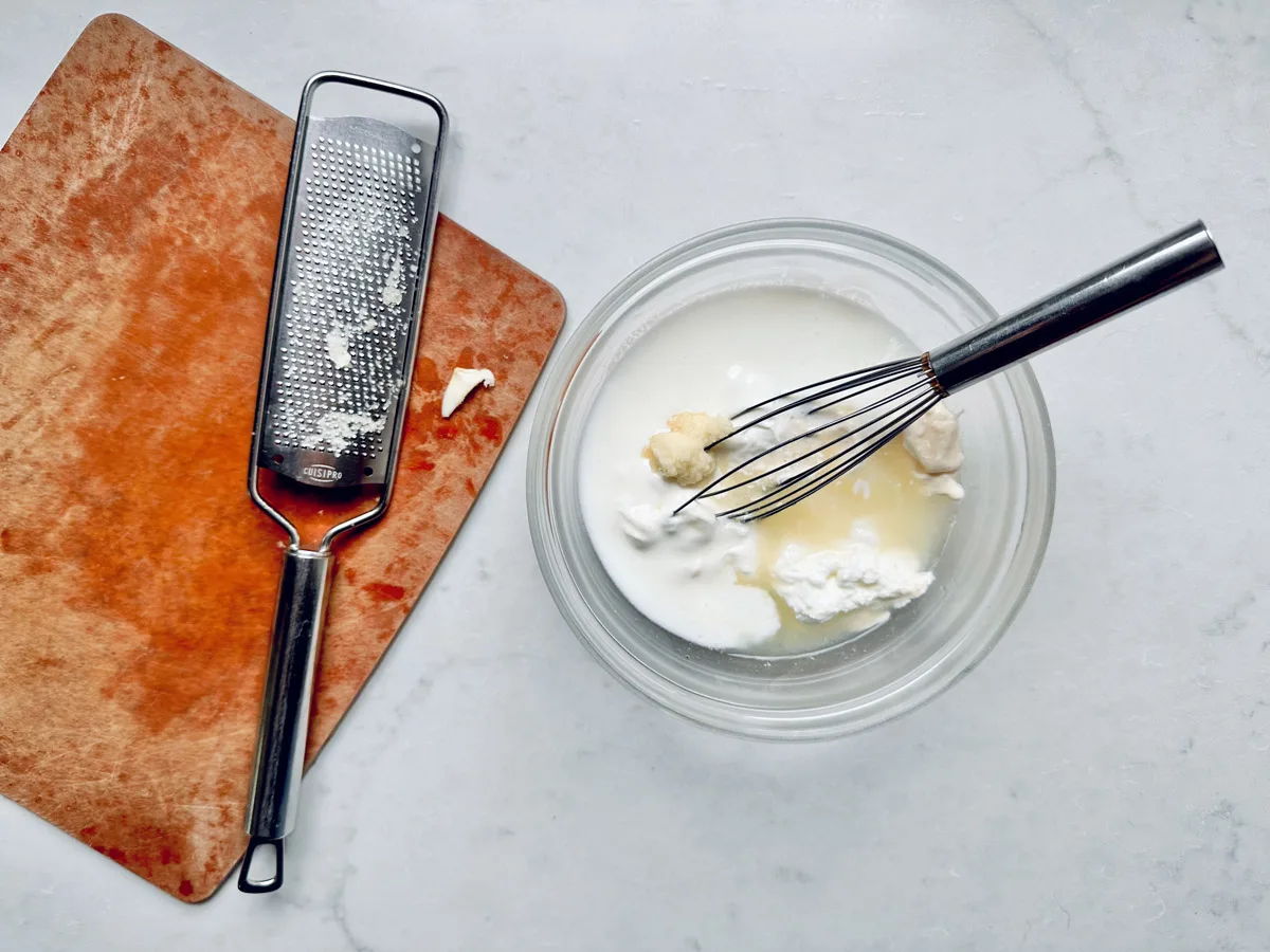 A glass bowl with milk, a chunk of PepperJack Cheese, and a whisk sits on a white surface next to a grater and an orange cutting board with cheese residue.