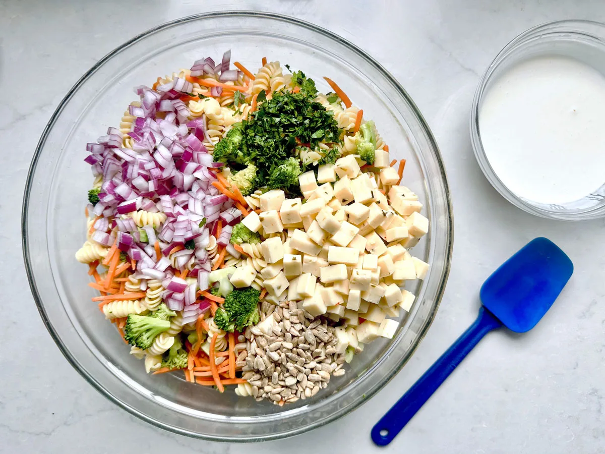 A glass bowl holds un-mixed Pasta Salad with Broccoli ingredients: diced red onion, parsley, carrots, rotini pasta, cubes of PepperJack Cheese, shredded carrots, and sunflower seeds. A bowl of white dressing and a blue spatula sit nearby.
