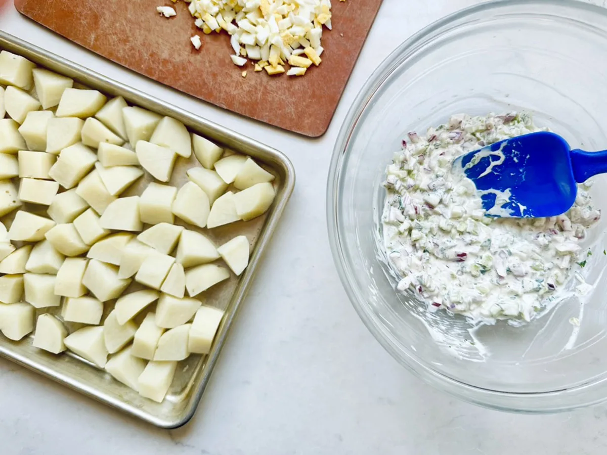 Chopped potatoes for All-American Russet Potato Salad rest on a baking sheet, diced hard-boiled eggs sit on a cutting board, and a bowl of creamy salad mixture is stirred with a blue spatula on a white surface.