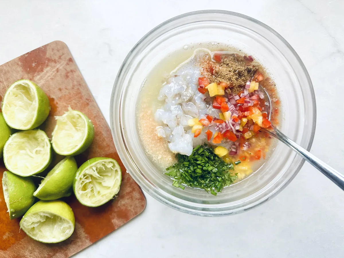 A glass bowl with shrimp, chopped herbs, diced red peppers, spices, and liquid ingredients, with a spoon mixing them. Next to it, a cutting board holds several squeezed lime halves.