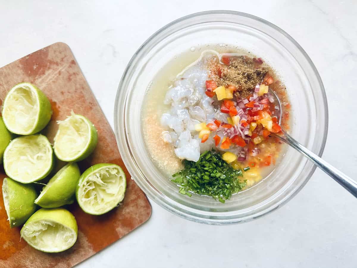 A glass bowl with shrimp, chopped herbs, diced red peppers, spices, and liquid ingredients, with a spoon mixing them. Next to it, a cutting board holds several squeezed lime halves.