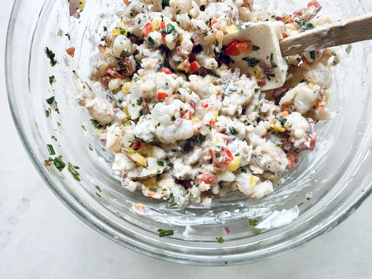 A glass bowl filled with a creamy shrimp salad mixed with chopped red and yellow bell peppers and fresh herbs, being stirred with a white spatula.