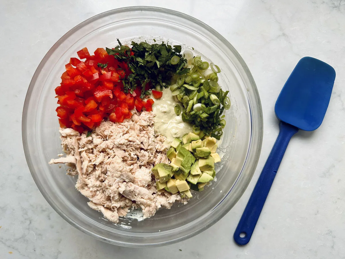 A glass bowl holds Spicy Chicken Salad with Chipotle and Avocado, featuring chopped red bell pepper, cilantro, green onions, avocado cubes, and shredded chicken in sections. A blue spatula rests on the right atop a light countertop.