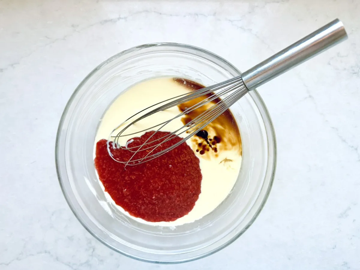 A metal whisk rests in a glass bowl containing heavy cream, bright red tomato sauce, Worcestershire sauce, and black pepper&mdash;ingredients mingling atop a white marble surface reminiscent of crafting Strawberry-Balsamic Ice Cream.