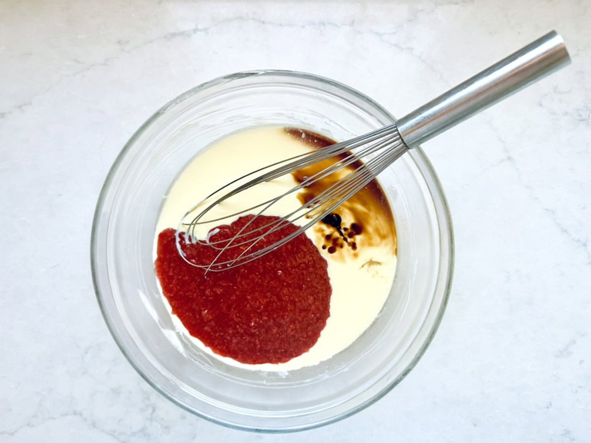 A metal whisk rests in a glass bowl containing heavy cream, bright red tomato sauce, Worcestershire sauce, and black pepper—ingredients mingling atop a white marble surface reminiscent of crafting Strawberry-Balsamic Ice Cream.