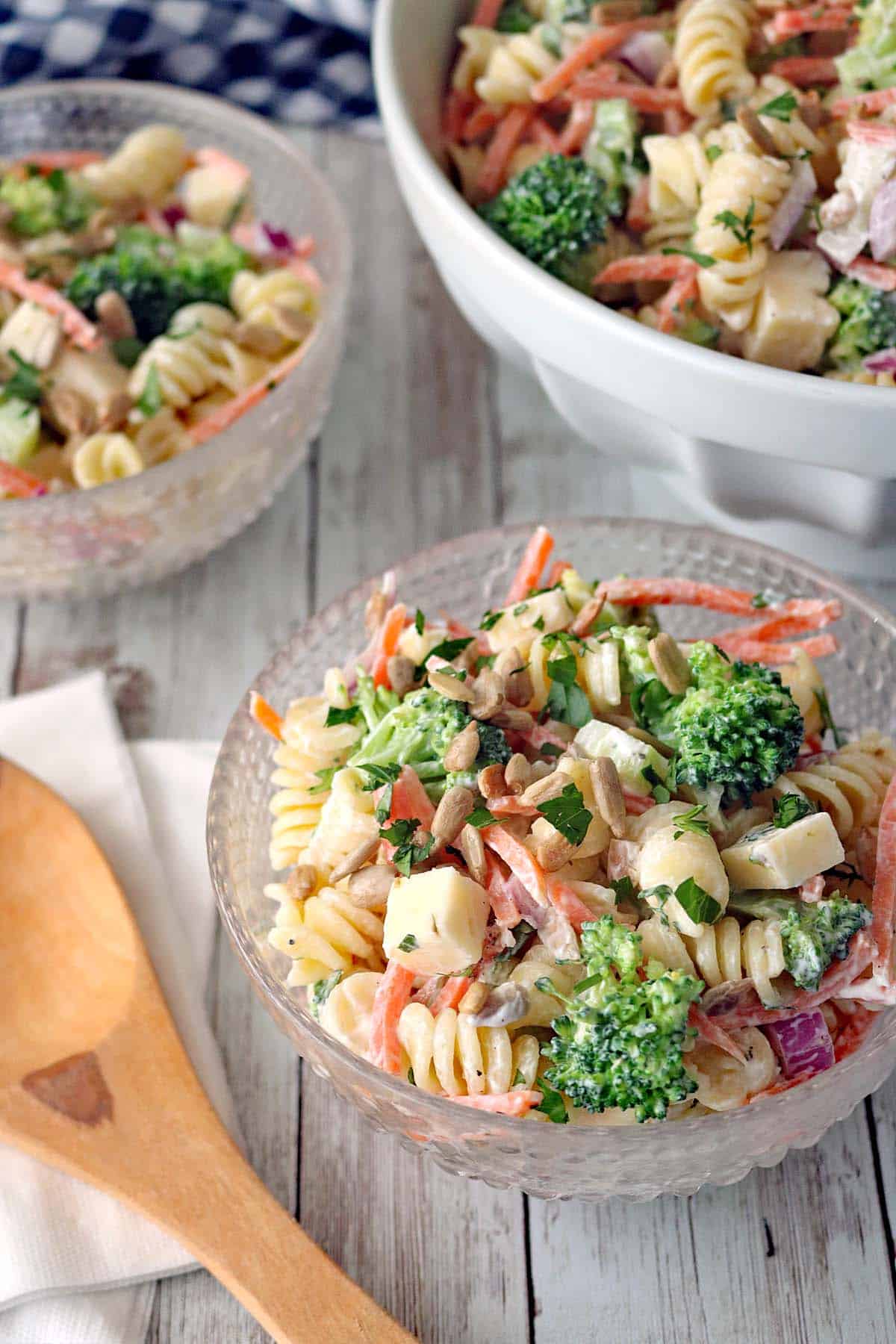 A close-up of Pasta Salad with Broccoli in glass bowls, featuring rotini pasta, carrots, red onion, PepperJack cheese cubes, sunflower seeds, and herbs in a creamy dressing. A wooden spoon and napkin are nearby.