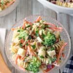 A glass bowl of Pasta Salad with Broccoli, carrots, and PepperJack cheese cubes is lightly coated in a creamy dressing. A wooden spoon and a napkin sit nearby on the rustic wooden table.