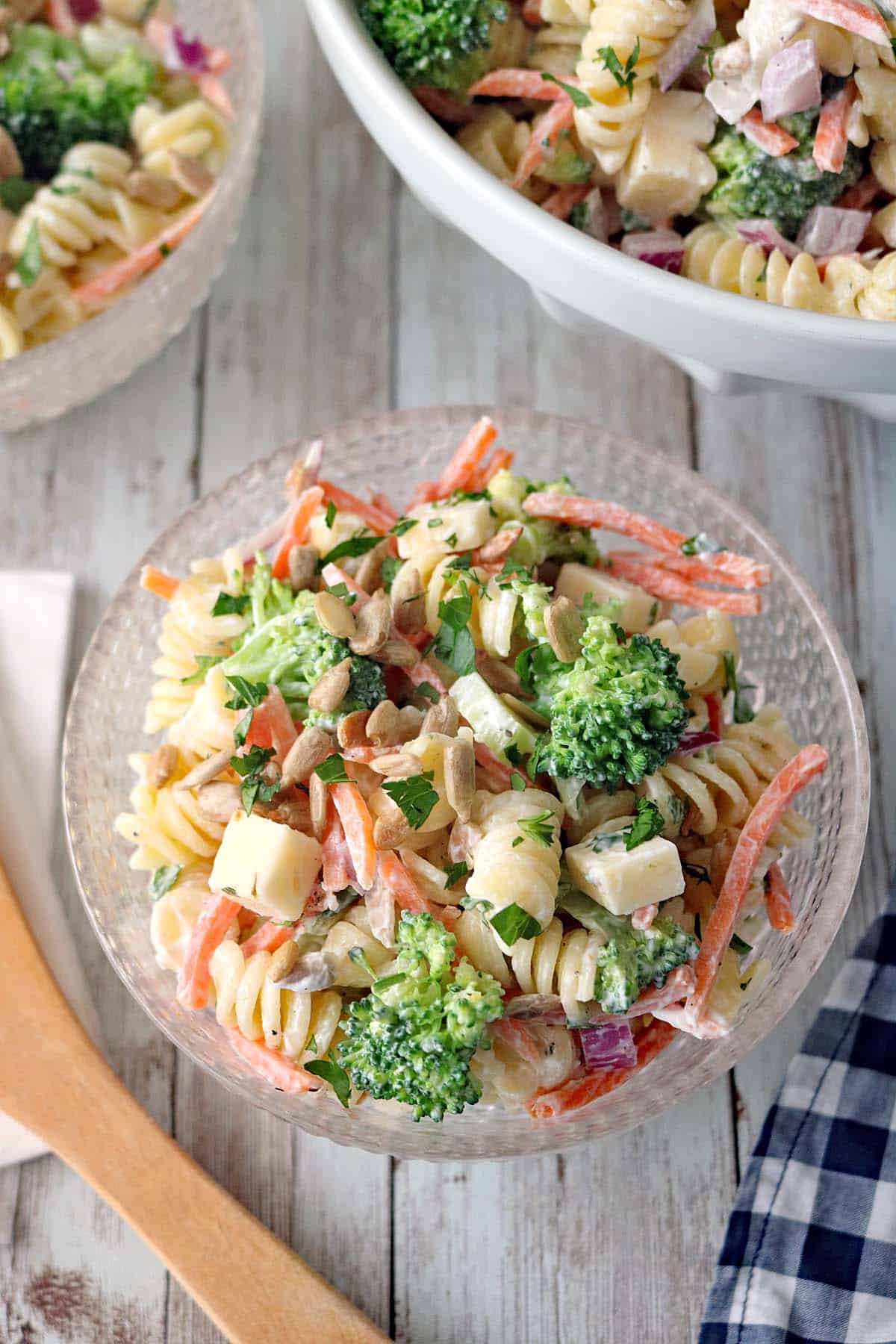 A glass bowl of Pasta Salad with Broccoli, carrots, and PepperJack cheese cubes is lightly coated in a creamy dressing. A wooden spoon and a napkin sit nearby on the rustic wooden table.