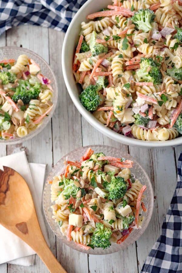 A bowl and two small dishes of Pasta Salad with Broccoli, Carrots, red onion, and PepperJack Cheese cubes, all tossed in dressing on a wooden table next to a wooden spoon and checkered cloth.