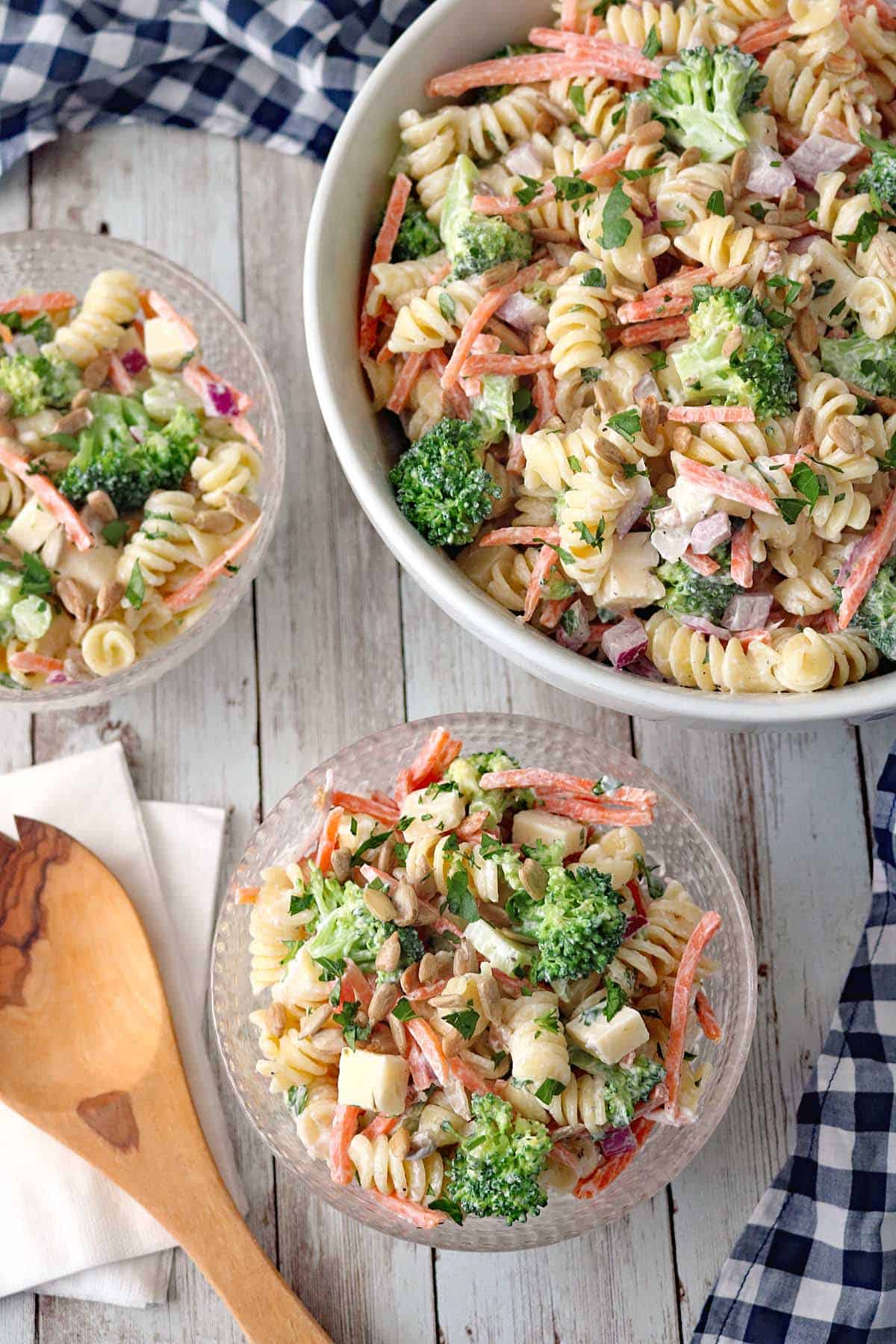 A bowl and two small dishes of Pasta Salad with Broccoli, Carrots, red onion, and PepperJack Cheese cubes, all tossed in dressing on a wooden table next to a wooden spoon and checkered cloth.