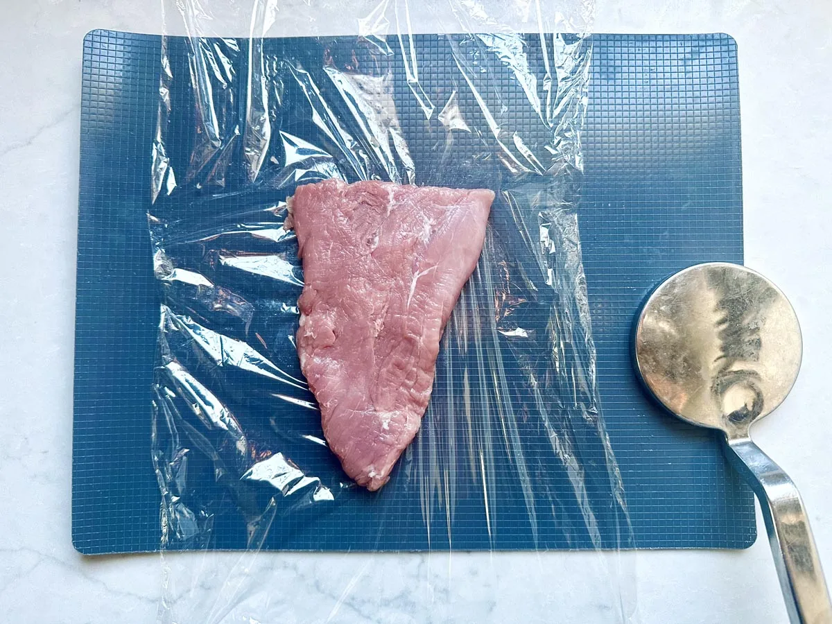 A raw piece of meat sits on a blue cutting board, partially covered with plastic wrap. A metal meat tenderizer is placed to the right of the meat.