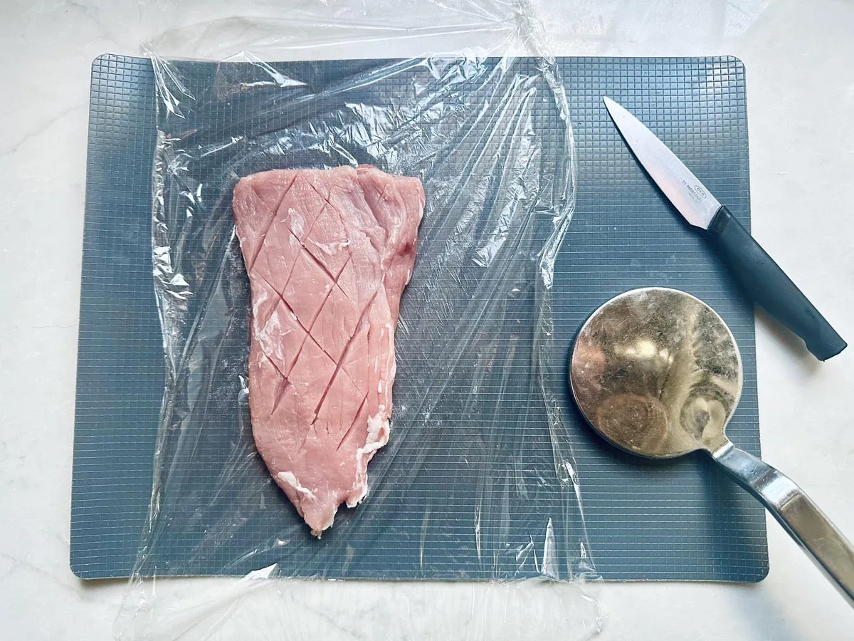 A raw piece of meat with crisscross cuts lies on a cutting board, covered partly by plastic wrap. A metal mallet and a knife are placed next to the meat.