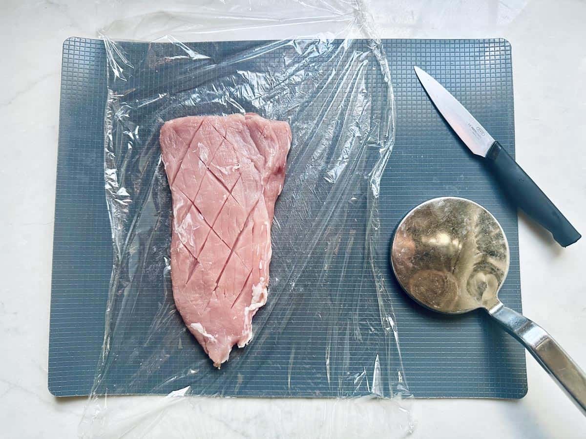 A raw piece of meat with crisscross cuts lies on a cutting board, covered partly by plastic wrap. A metal mallet and a knife are placed next to the meat.