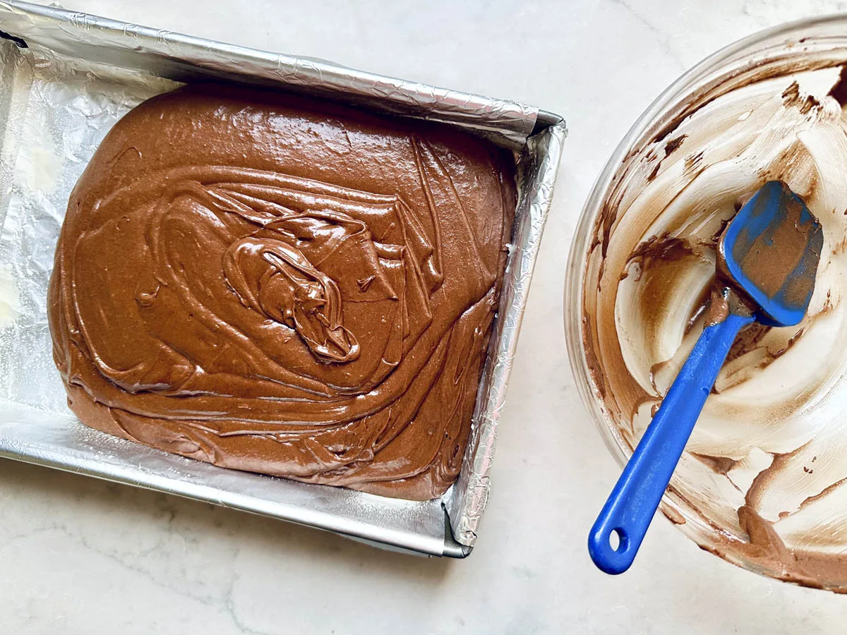 A baking pan lined with foil filled with Chocolate Chipotle Brownies batter sits beside a glass bowl with chocolate residue and a blue spatula on a light countertop.