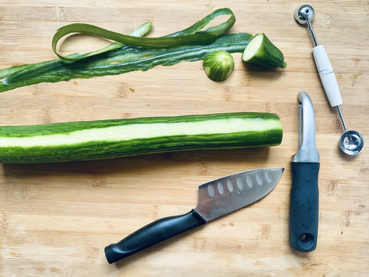 A partially peeled cucumber on a wooden board with a knife, peeler, melon baller, cucumber slices, and cucumber peel next to it.