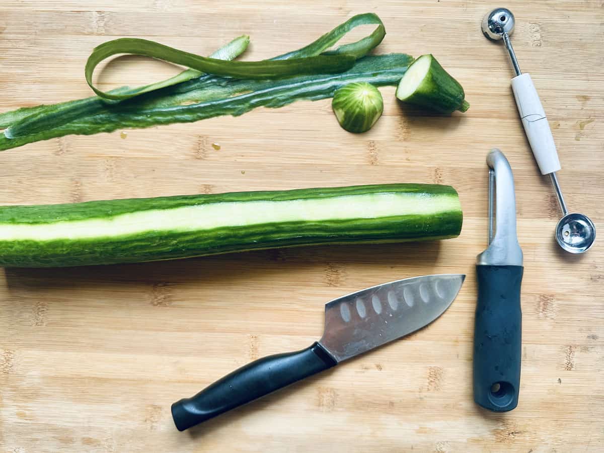A partially peeled cucumber on a wooden board with a knife, peeler, melon baller, cucumber slices, and cucumber peel next to it.