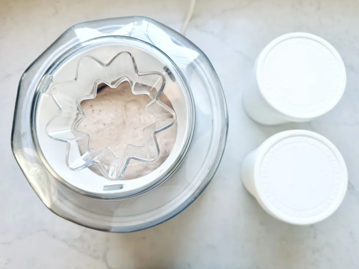 Top view of an ice cream maker churning creamy Strawberry-Balsamic Ice Cream, next to two white lidded containers on a light countertop.