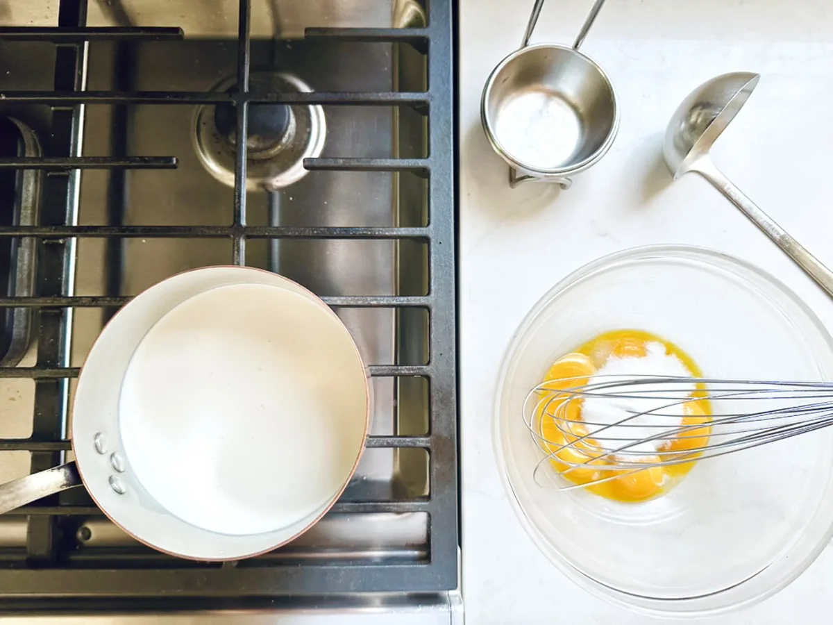 Overhead view of a stovetop with a saucepan, beside a counter holding ingredients and tools for Strawberry-Balsamic Ice Cream: a clear bowl with egg yolks and sugar, a whisk, ladle, and two measuring cups.