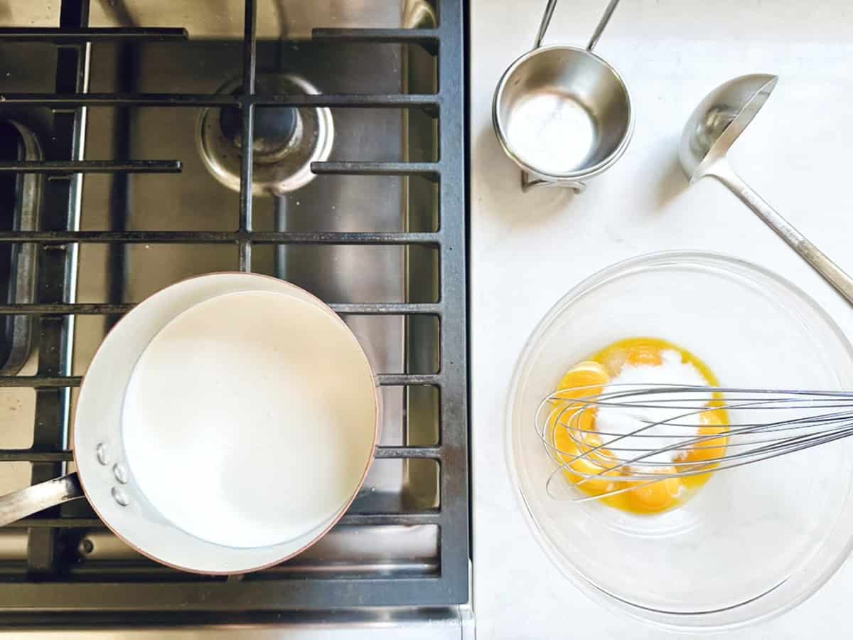 Overhead view of a stovetop with a saucepan, beside a counter holding ingredients and tools for Strawberry-Balsamic Ice Cream: a clear bowl with egg yolks and sugar, a whisk, ladle, and two measuring cups.