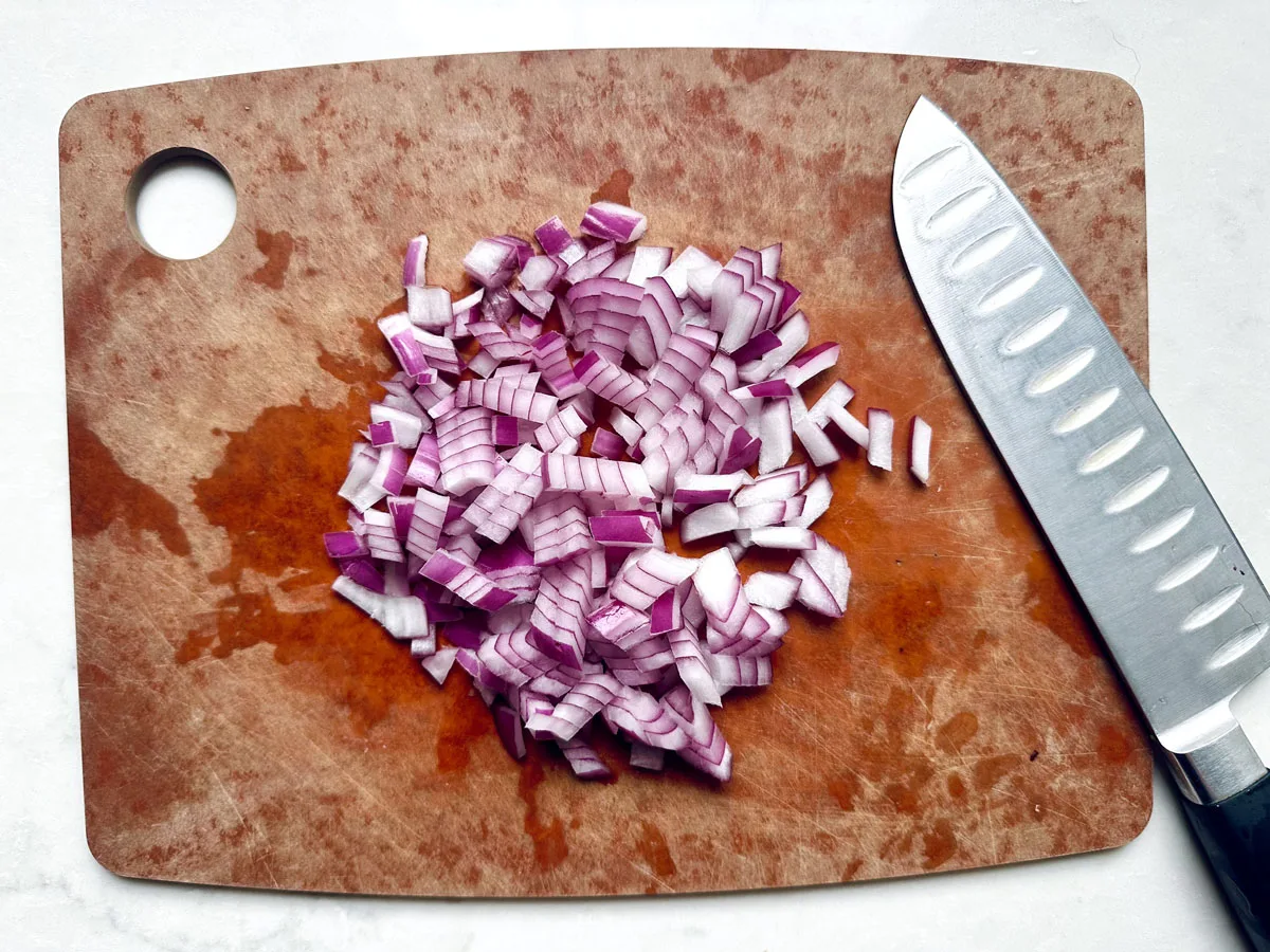 Finely diced red onion on a wooden cutting board with a chef’s knife beside it; the board shows juice stains—perfect prep for Pasta Salad with Broccoli, Carrots, and PepperJack Cheese.