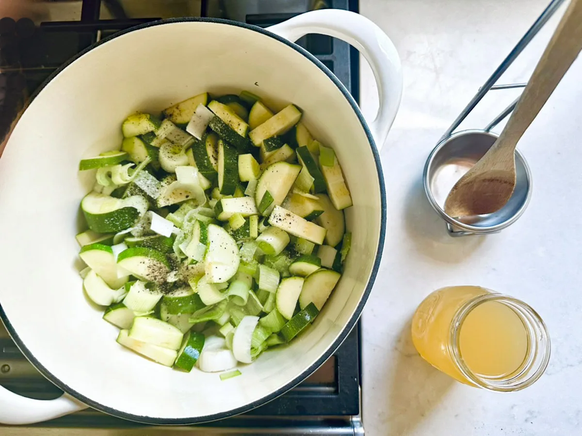 A white pot on a stove contains chopped zucchini and onions for Zucchini Soup with Dill and Greek Yogurt, seasoned with black pepper. Nearby sit a jar of broth and a metal cup holding a wooden spoon and measuring spoon.