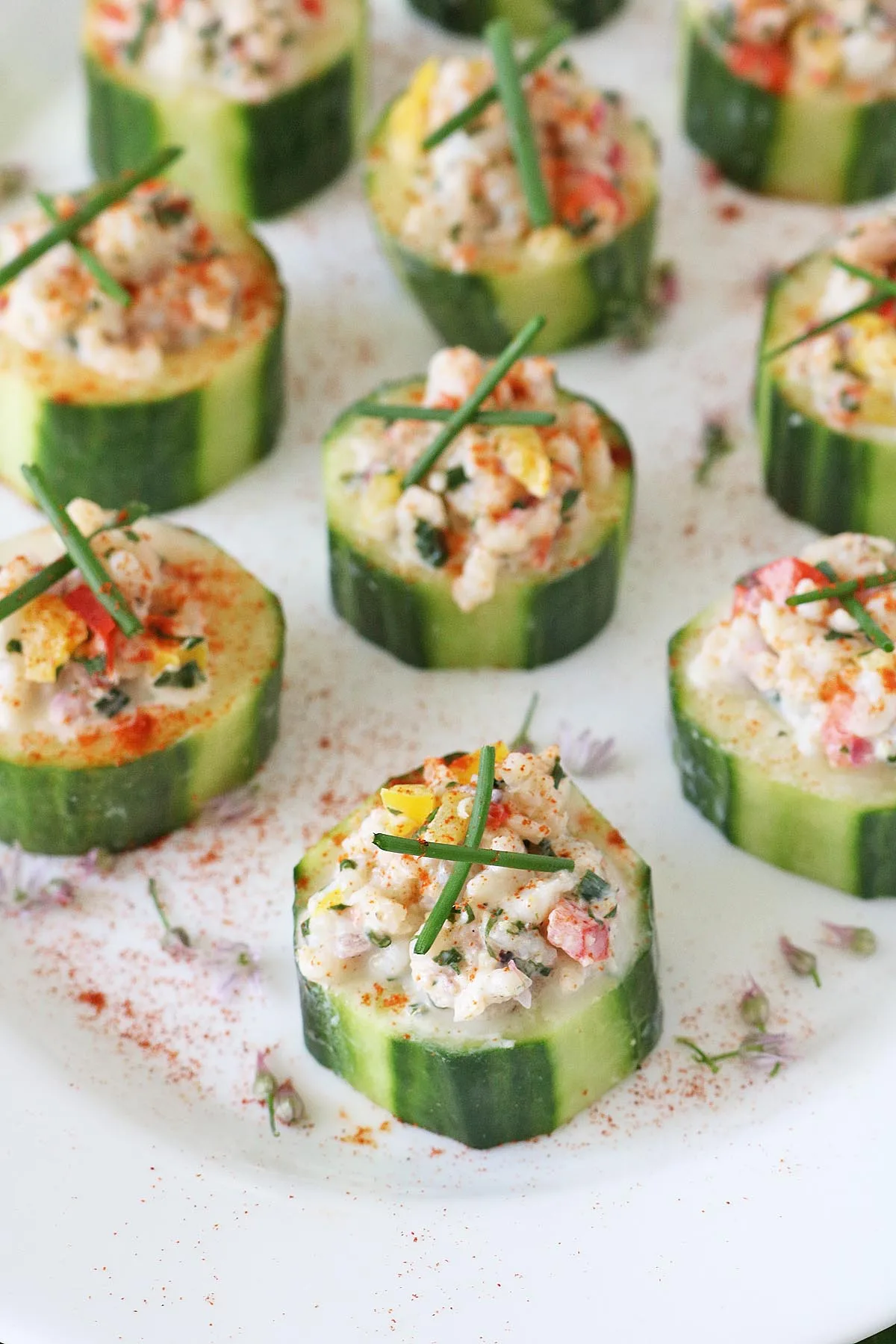 Cucumber slices topped with a creamy seafood filling, garnished with herbs and thin chive sprigs, arranged neatly on a white plate with a sprinkle of paprika.