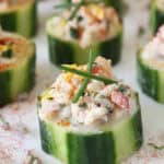 Close-up of cucumber slices topped with a creamy seafood salad mixture, garnished with chives and a sprinkle of paprika, arranged on a white surface.