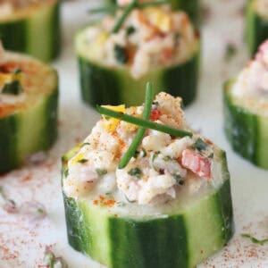 Close-up of cucumber slices topped with a creamy seafood salad mixture, garnished with chives and a sprinkle of paprika, arranged on a white surface.