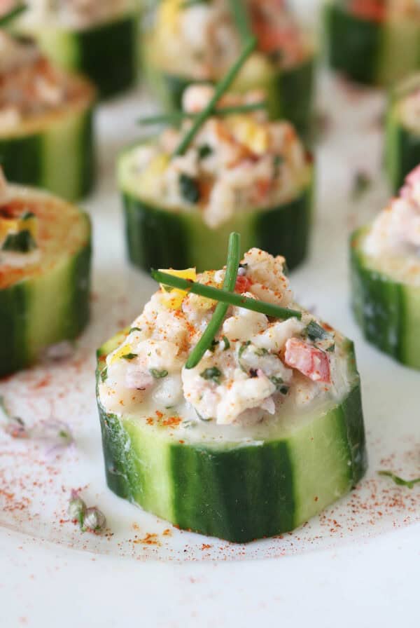 Close-up of cucumber slices topped with a creamy seafood salad mixture, garnished with chives and a sprinkle of paprika, arranged on a white surface.
