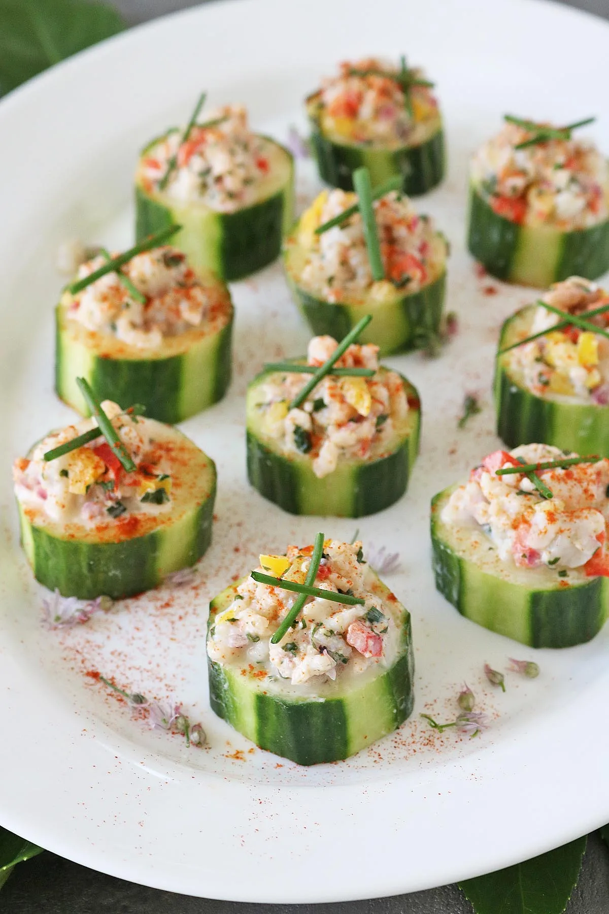 Sliced cucumber rounds topped with a creamy seafood and vegetable mixture, garnished with chives and sprinkled with paprika, arranged neatly on a white plate.