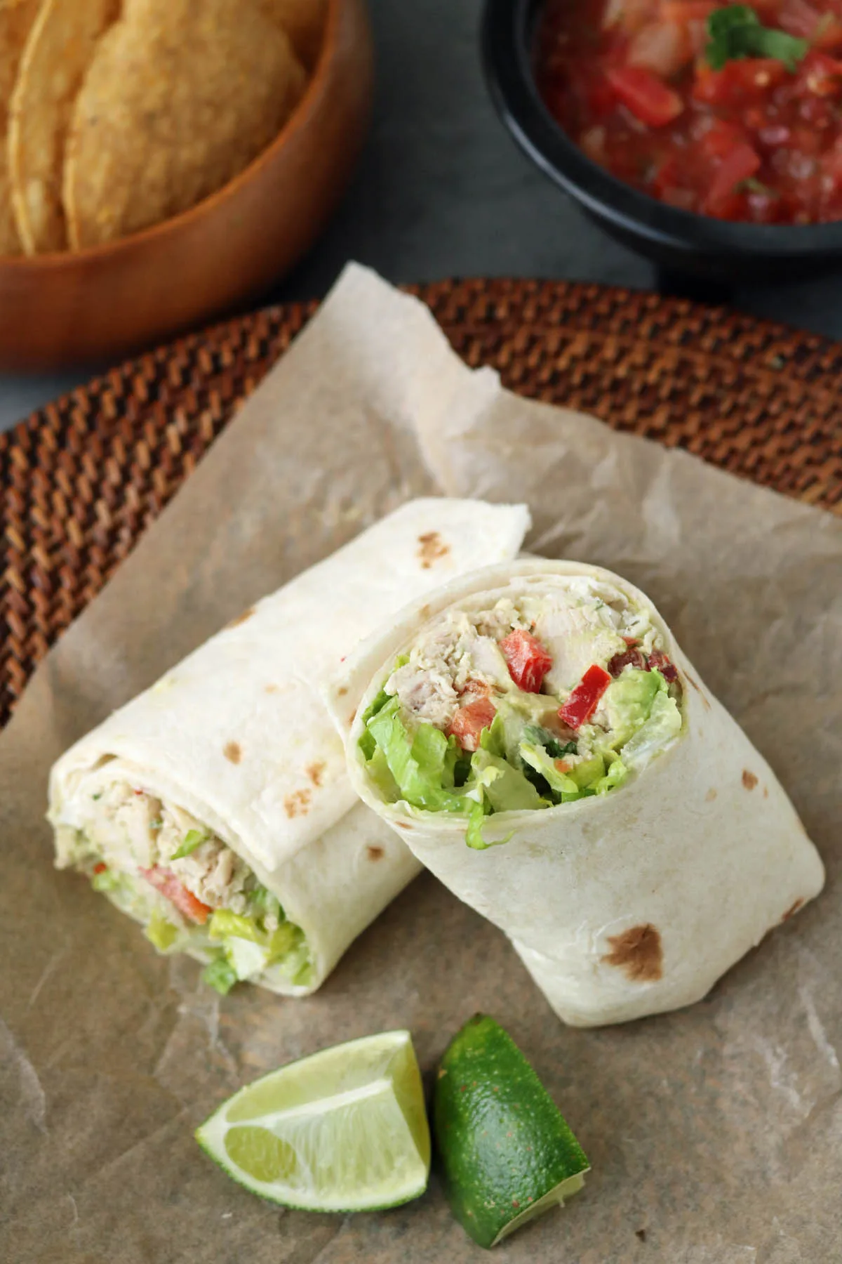 A Spicy Chicken Salad with Chipotle and Avocado wrap, cut in half and filled with lettuce and red bell peppers, sits on parchment paper with lime wedges. Tortilla chips in a wooden bowl and a bowl of salsa are in the background.