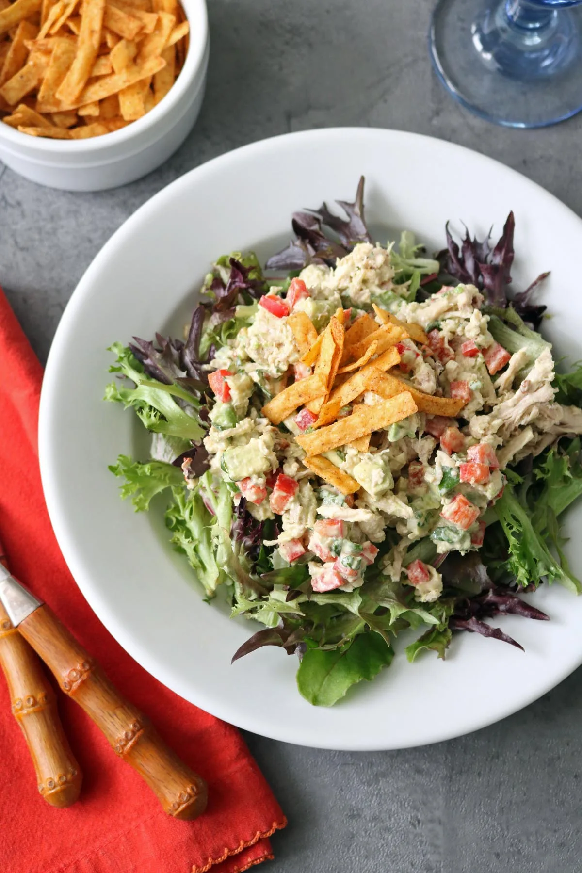A white plate of Spicy Chicken Salad with Chipotle and Avocado, featuring diced red bell peppers and creamy dressing on leafy greens, topped with crispy tortilla strips. Extra strips and bamboo-handled cutlery sit nearby on a gray surface with a red napkin.